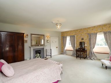 A bedroom with a large bed and pillows a wardrobe a fireplace a dressing table two windows with curtains and a patterned yellow wallpaper at Craster Tower Penthouse in Craster