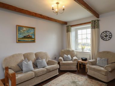A living room with beige sofas cushions a small wooden table a window with curtains a wall clock and a framed painting at No2 Budle Bay Cottage Bamburgh