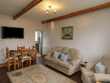 A living room with a dining table and chairs a sofa and armchair a wall-mounted tv and a painting at No2 Budle Bay Cottage in Bamburgh