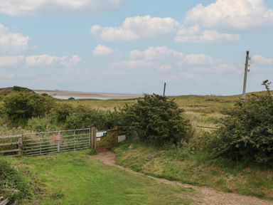 A grassy field with bushes and two gates leading to open land and a sandy area in the distance at No2 Budle Bay Cottage in Bamburgh