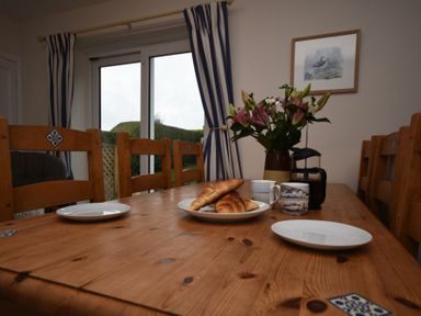 A dining table with plates croissants mugs flowers and a french press by window with curtains at Inveray in Seahouses