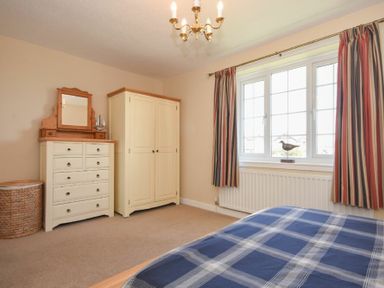 A bedroom with a bed with blue checkered bedding a white wardrobe a white chest of drawers with a mirror a wicker basket striped curtains and a window at Inveray in Seahouses