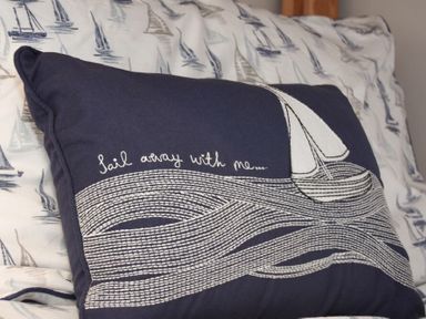 A navy pillow with a sailboat and the words sail away with me on a bed with sailboat printed bedding at Inveray in Seahouses