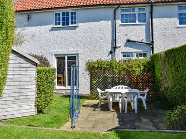 A backyard with a white plastic table and chairs on a paved area next to a wooden shed and a white house with a tiled roof at Inveray in Seahouses