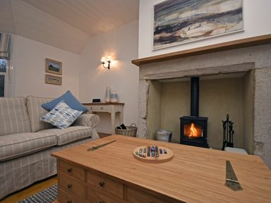 A living room with a striped sofa and cushions a wooden table with a game a fireplace with a fire and painting above at Periwinkle in Bamburgh