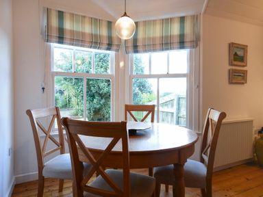 A dining area with a round wooden table and four chairs by two large windows with striped blinds at Periwinkle in Bamburgh