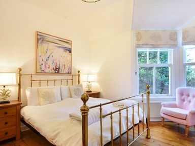 A bedroom with a brass bed frame two wooden bedside tables with lamps a pink armchair and two windows at Periwinkle in Bamburgh