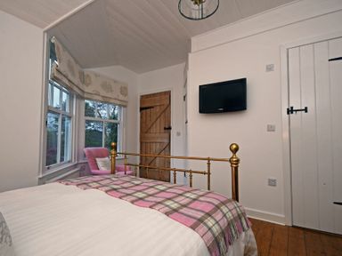 A bedroom with a brass bed a pink chair near windows a wooden door and a wall-mounted TV at Periwinkle in Bamburgh