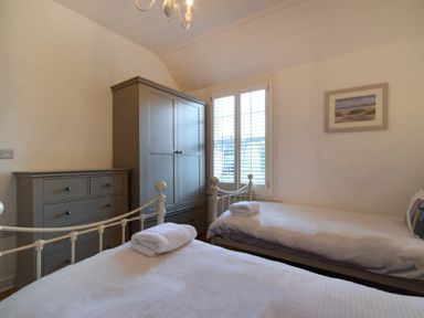 A bedroom with two single beds white linen a wardrobe a chest of drawers and a window with shutters at Periwinkle in Bamburgh