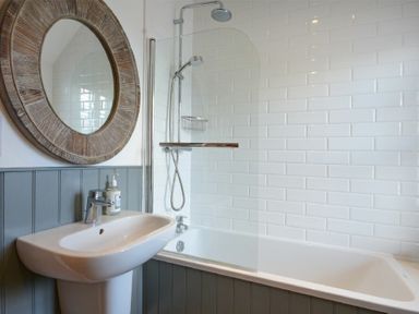 A bathroom with a bathtub shower combination a round wooden framed mirror a white sink and soap dispenser at Periwinkle in Bamburgh