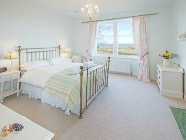 A bedroom with a brass bed frame two bedside tables with lamps a window with curtains and a white dresser with flowers at The Villas No3 Embleton