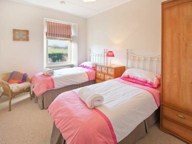 A bedroom with two single beds with pink and white bedding a wooden wardrobe and a wicker chair at The Villas No3 in Embleton
