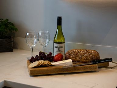 A wooden cutting board with a loaf of seeded bread a knife cheese grapes crackers two tomatoes a bottle of white wine and two wine glasses on a kitchen counter at Castle View Bamburgh