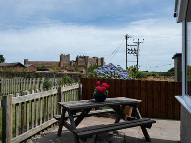 A wooden picnic table with a pot of red flowers on a patio with a wooden fence and a view of a stone castle in the background at Castle View Bamburgh