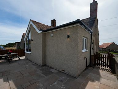Exterior view of a house with a paved patio and wooden picnic table with flowers at Castle View Bamburgh
