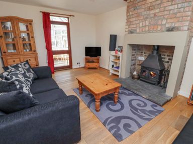 A living room with a black sofa and patterned cushions a wooden coffee table on a patterned rug a wood burning stove in a brick fireplace a wooden cabinet a television on a wooden stand a white shelving unit and a door with a red curtain at St Marys Cottages No1 in Low Newton-by-the-Sea