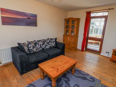 A living room with a black sofa with patterned cushions a wooden coffee table a wooden cabinet and a door leading to a conservatory at St Marys Cottages No1 in Low Newton-by-the-Sea