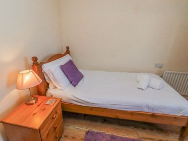 A bedroom with a single wooden bed with white bedding and a purple cushion next to a wooden nightstand with a lamp and coasters at St Marys Cottages No1 in Low Newton-by-the-Sea