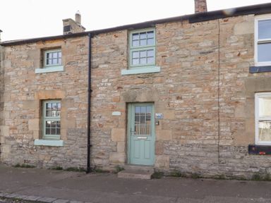 A stone exterior of a house with a green door and three green framed windows at Hares Form in Seahouses