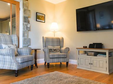 A living room with two armchairs beside a small table a floor lamp and a cabinet with a TV on the wall at The Pepper Pot in Warkworth