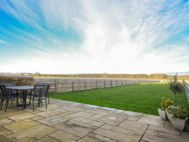 A patio with a table and chairs next to a grassy yard with a fence and plants at The Pepper Pot in Warkworth