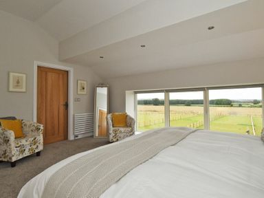 A bedroom with a bed two armchairs a mirror and large windows overlooking fields at The Pepper Pot in Warkworth
