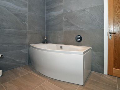 A bathroom with a white bathtub grey tiled walls and floor and a wooden door at The Pepper Pot in Warkworth