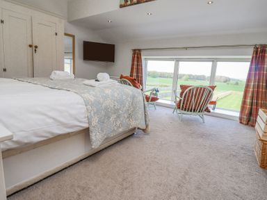 A bedroom with a bed folded towels a wall mounted tv two armchairs by a large window with curtains and a view of greenery at The Mustard Pot in Acklington