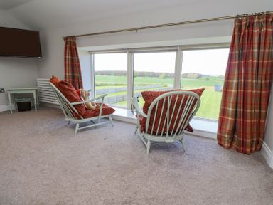 Two chairs with red cushions by a large window with a view of fields a wall-mounted TV and a small table in a carpeted room at The Mustard Pot in Acklington