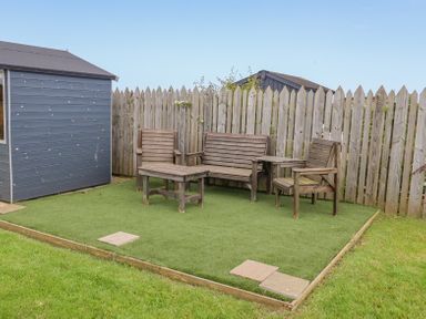 An outdoor seating area with wooden chairs, a bench, a coffee table, and a blue shed surrounded by a wooden picket fence at The Mustard Pot in Acklington