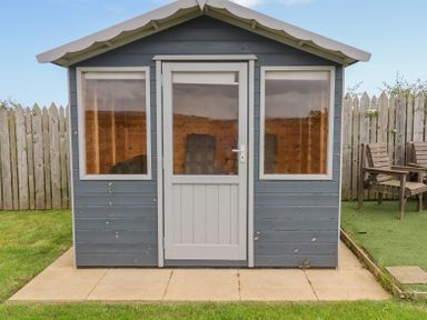 A wooden garden shed with glass windows and door on a stone patio with wooden chairs and a wooden fence in the background at The Mustard Pot in Acklington