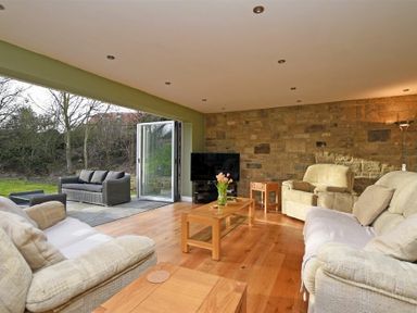 A living room with beige sofas wooden tables and a television with an open door leading to an outdoor patio at Quarry Haven in Bamburgh
