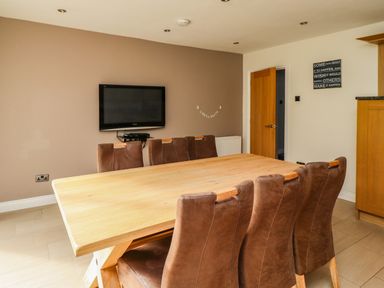 A dining room with a wooden table and brown chairs a wall mounted TV and a door at Quarry Haven in Bamburgh