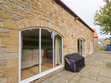 A stone wall with arched glass doors and a covered grill on a stone patio at Quarry Haven in Bamburgh