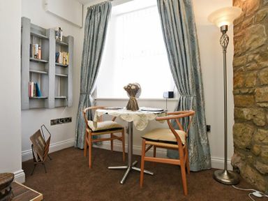 A small dining area with two chairs a round table with a tablecloth and flowers by a window with curtains and a floor lamp near a stone wall at Sea Air in Amble