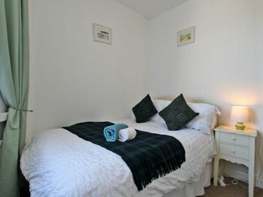 A bedroom with a bed with white bedding and two dark pillows two rolled towels on a blanket a bedside table with a lamp and green curtains at Sea Air in Amble