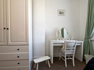 A bedroom corner with a white wardrobe a small wooden stool a white dressing table with a triple mirror and a white chair with cushions green curtains and a laundry basket at Sea Air in Amble