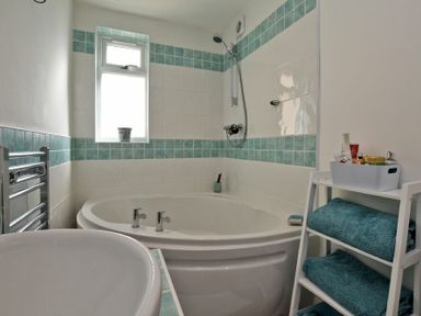 A bathroom with a corner bathtub shower towel rack and a shelving unit with folded towels and toiletries at Sea Air in Amble