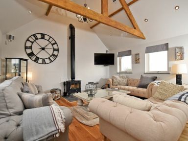 A living room with beige and gray sofas a wooden coffee table a black wood stove a large wall clock and exposed wooden ceiling beams at The Hemmel (Beadnell) in Beadnell