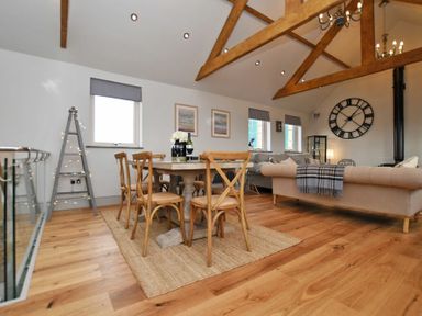 A dining area with wooden chairs and a table on a rug next to a living room with sofas and a large clock on the wall at The Hemmel (Beadnell) in Beadnell