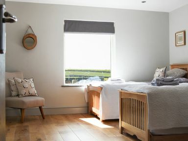 A bedroom with two wooden beds a beige chair with cushions a window and a round wall mirror at The Hemmel in Beadnell