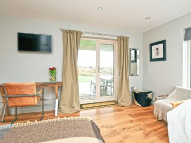 A room with a wall-mounted TV a wooden desk with a chair a glass door with curtains and a chair with a throw at The Hemmel Beadnell