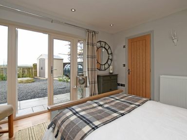 A bedroom with a bed covered with a plaid blanket a wooden door a green cabinet with a round mirror and glass doors leading outside at The Hemmel Beadnell