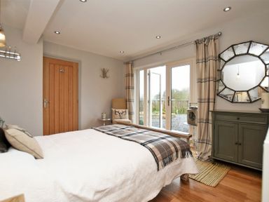 A bedroom with a wooden door bed with cushions and blanket a chair by glass doors and a cabinet with a mirror at The Hemmel (Beadnell) in Beadnell