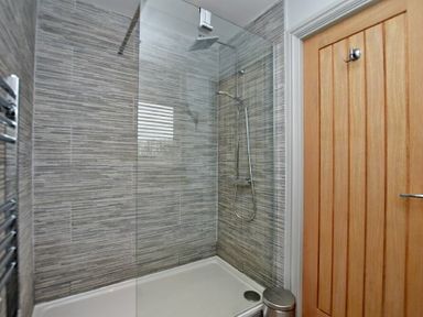 A shower area with gray tiled walls a glass shower screen a rainfall showerhead a wooden door and a small metal trash bin at The Hemmel Beadnell