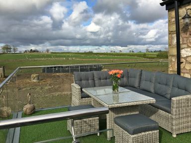 An outdoor seating area with a corner sofa table and stool on artificial grass overlooking a green field at The Hemmel (Beadnell) in Beadnell