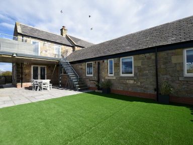A stone house with a patio table chairs stairs and artificial grass at The Hemmel (Beadnell) in Beadnell