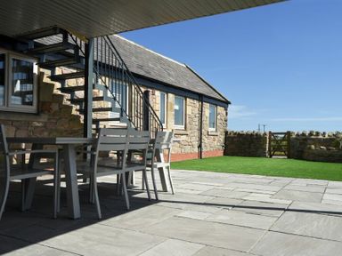 An outdoor patio with a table and chairs under a roof and a stone building with stairs and a grassy area at The Hemmel Beadnell