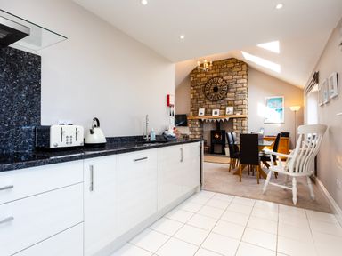 A kitchen with white cabinets and black countertops next to a dining area with a stone fireplace at Harbour Lights House in Seahouses