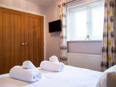 A bedroom with a bed holding rolled towels a window with patterned curtains a wall mounted tv and wooden closet doors at Harbour Lights House in Seahouses